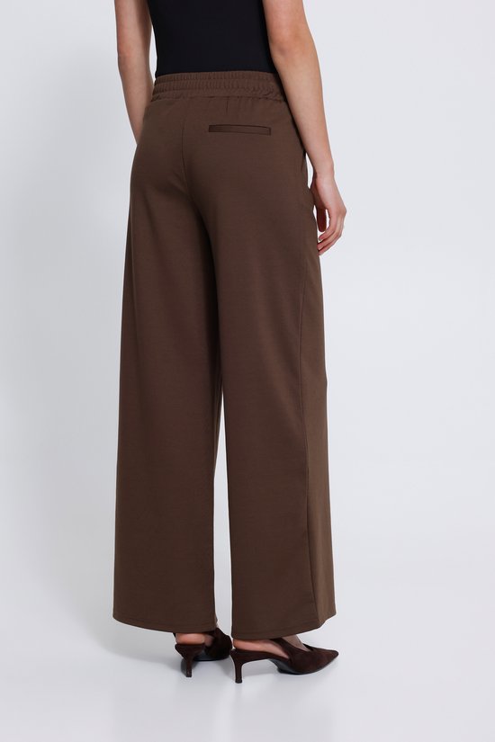 Achterkant van een model in een bruine wijde broek met een elastische tailleband en één zak aan de achterkant.
