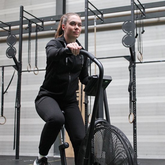 Vrouw met paardenstaart in een zwarte hoodie en zwarte broek, op een hometrainer.