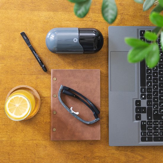 Een AYO-lichttherapiebril ligt op een bruine notitieboekje naast een laptop, glas jus d'orange en een zwarte pen op een houten tafel.