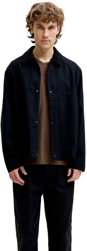 Man met krullend haar draagt een zwarte jas over een bruin T-shirt en zwarte broek.