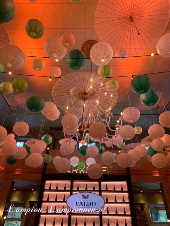 Een interieur met veel witte en groene lampionnen, parasols en een kroonluchter aan het plafond. Oranje gloed.