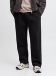 Model draagt zwarte wide-leg joggingbroek, beige t-shirt, getextureerde jas en sneakers.