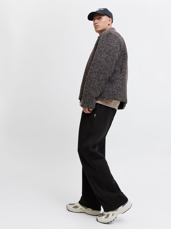 Zijaanzicht: model in zwarte wide-leg joggingbroek, beige t-shirt, getextureerde jas en pet.
