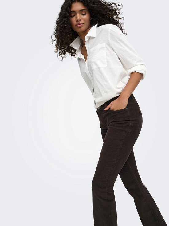 Model met krullend haar draagt een bruine corduroy flared broek en witte blouse, handen in de zakken.