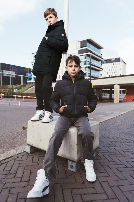 Twee jongens met winterjassen, een staat leunend tegen een paal en de ander zit met handen in de zakken.