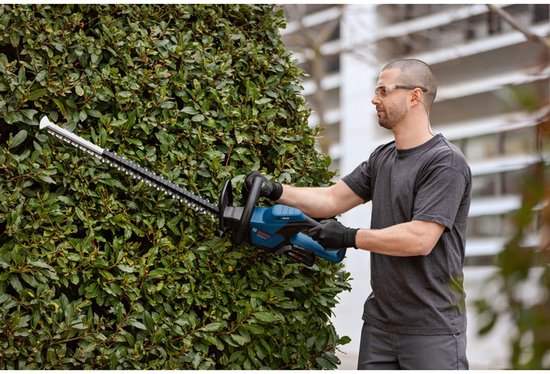 Man met blauwe en zwarte Bosch Professional heggenschaar snoeit een dichte groene heg.