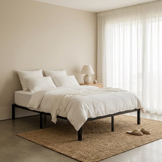 Zwart metalen bedframe met wit linnen beddengoed, nachtkastje en tafellamp. Een beige vloerkleed ligt onder het bed.