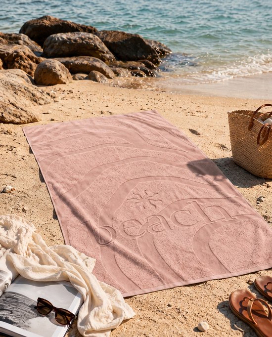 Zachtroze velours strandlaken met golvend patroon en het woord 'beach' in reliëf.