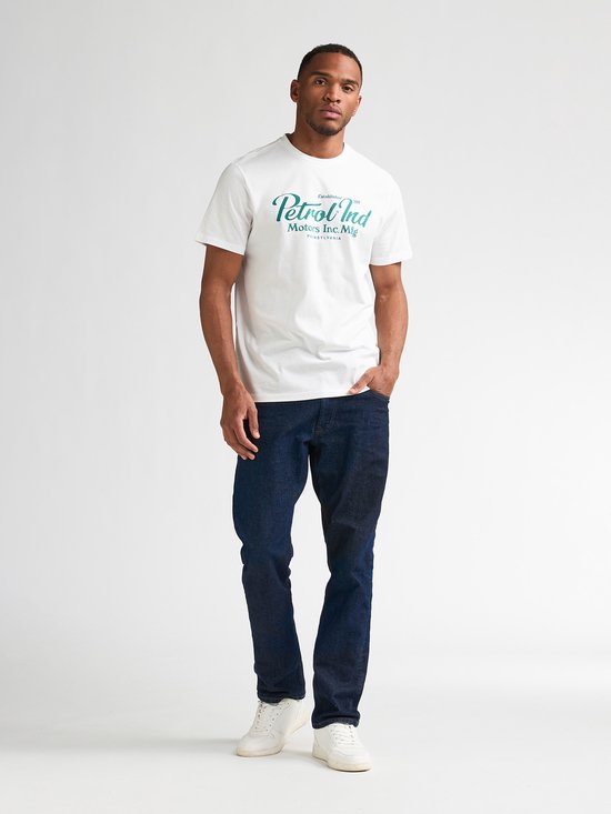 Man met donker haar, draagt wit T-shirt met groenblauwe print en spijkerbroek, volledig.