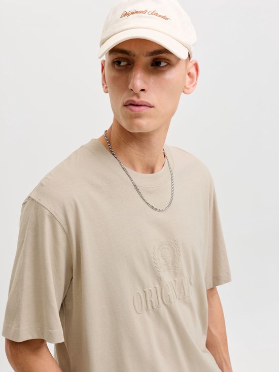 Man met beige T-shirt met "ORIGINALS" opdruk en beige pet, close-up.