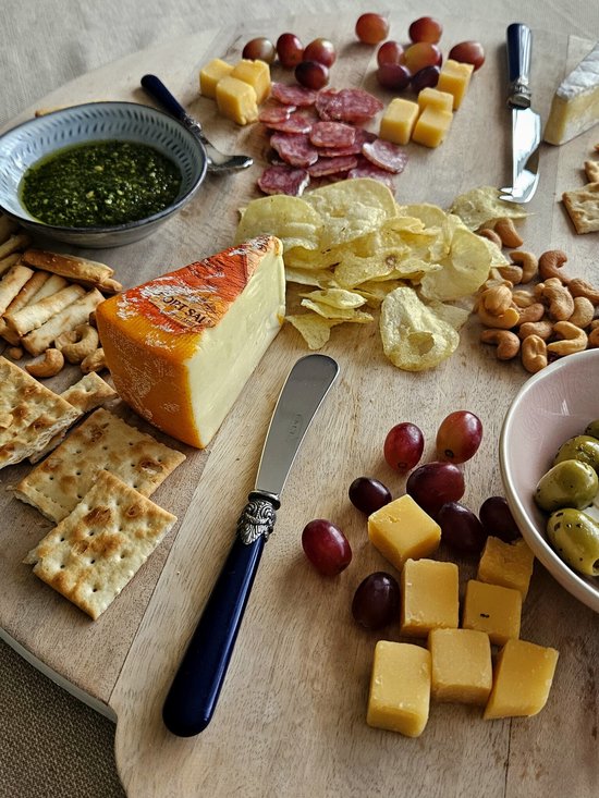 Een blauw botermesje met sierlijk zilverkleurig handvat op een kaasplank met kaas, worst, druiven, crackers en chips.