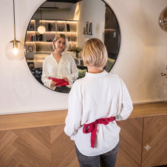 Vrouw in wit shirt knoopt een rood warmtekussen om haar middel, kijkt in spiegel.