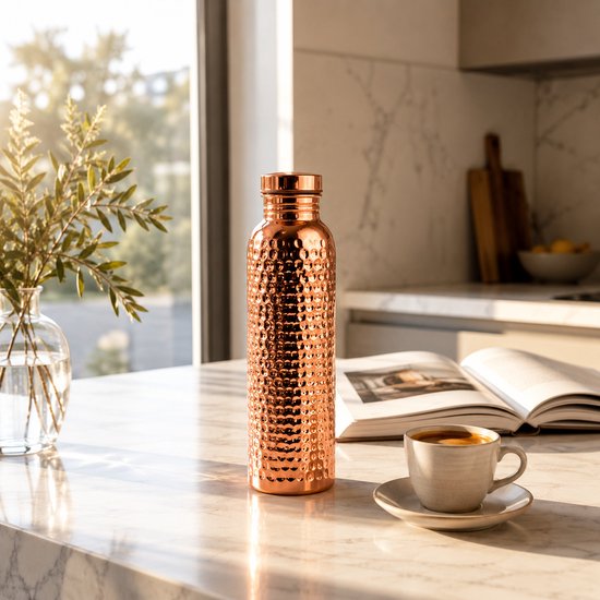 Koperen waterfles met hamerslag textuur op een marmeren aanrecht, naast een koffiekopje.