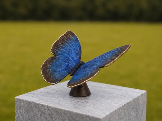 Papillon bleu en bronze aux bords dorés, posé sur un cube de pierre grise, sur fond vert.