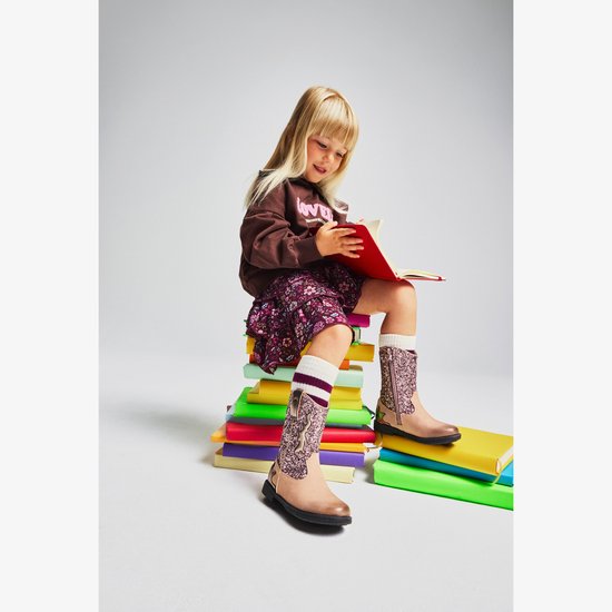 Meisje met blond haar, zittend op een stapel boeken, met bruine sweater en gebloemde rok.