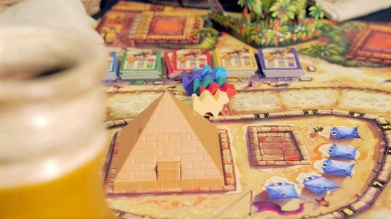 Een piramide-speelstuk, stenen munten, kamelen-speelstukken en kaarten op een spelbord.