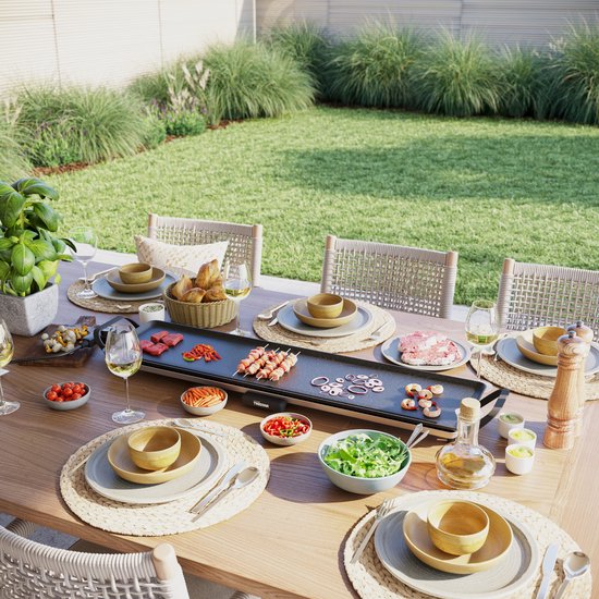 Table garnie de délices : steak grillé, crevettes, légumes et pain, sur un appareil à raclette dans un jardin.