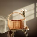 Panier rond en osier avec une anse, sur un tabouret avec un vase de fleurs et des ombres sur le mur.