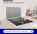 Zwarte inductiebeschermer met betonprint, bedekt met kruiden, knoflook, citroen en peperkorrels, naast een spatbord.
