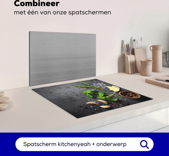 Zwarte inductiebeschermer met betonprint, bedekt met kruiden, knoflook, citroen en peperkorrels, naast een spatbord.