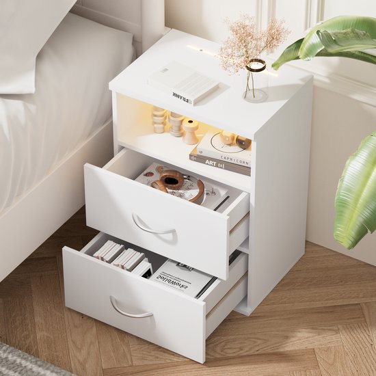 Wit nachtkastje met open vak, verlichting, decoratie, boeken en drie open lades met spullen.