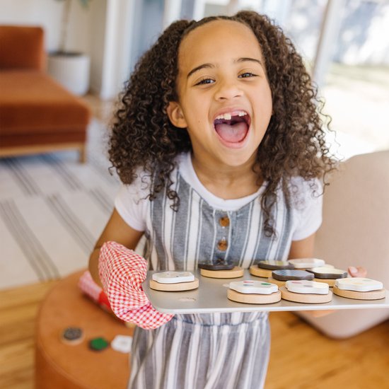 Lachend meisje met krullend haar, houdt bakplaat met houten koekjes met witte icing en gekleurde toppings vast. Ze draagt een rood-wit geblokt ovenwant.