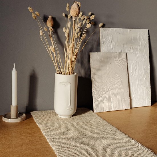 Een witte vaas met gezicht, gedroogde bloemen, kaars en twee witte textuurkunstwerken.