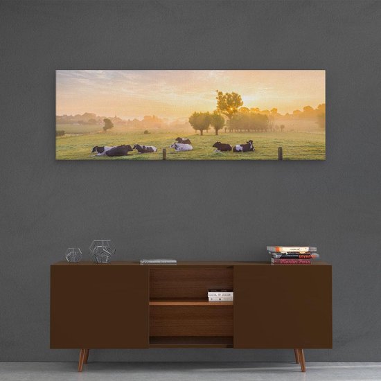 Een breed schilderij van koeien in een weiland bij zonsopgang, hangend boven een bruine dressoir met boeken.