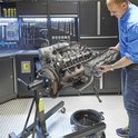 Homme en chemise bleue montant un moteur de voiture sur un support moteur dans un atelier.