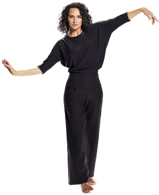 Model in een zwarte, wijde driekwartmouw top en zwarte broek met hoge taille, armen gespreid in een danshouding.