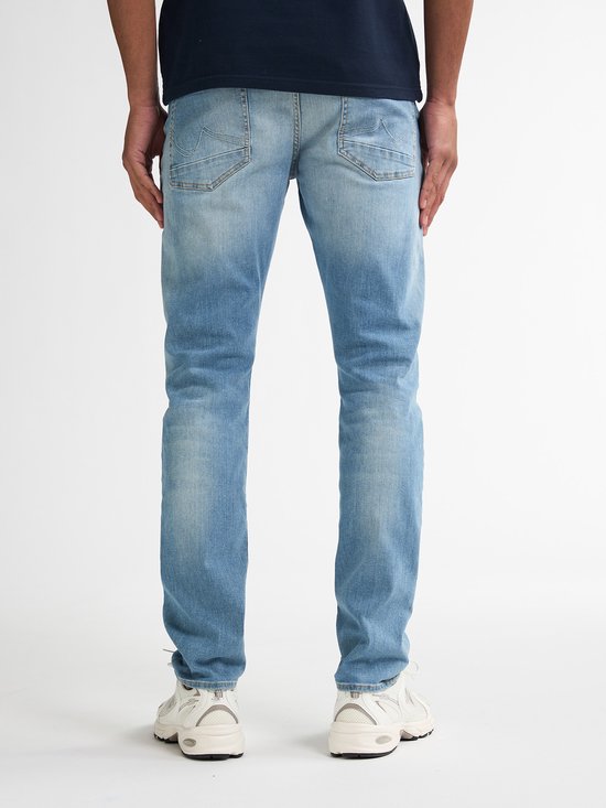 Man, van achteren gezien, draagt een marineblauwe polo en een lichtblauwe slim fit jeans met lichte wassing.