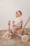 Baby in roze outfit op een roze driewieler met bloemenprint, naast een witte muur.