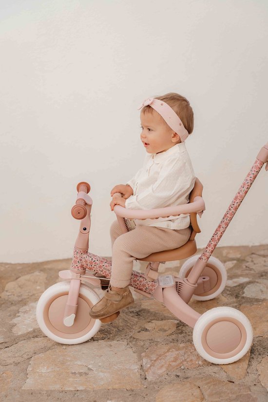 Baby in roze outfit op een roze driewieler met bloemenprint, naast een witte muur.