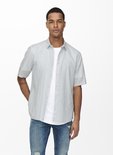Man met korte mouwen in lichtgrijs, open overhemd over een wit T-shirt en distressed jeans.