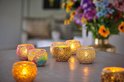 Meerdere mozaïek waxinelichthouders met LED kaarsjes erop, met een vaas met bloemen op de achtergrond.