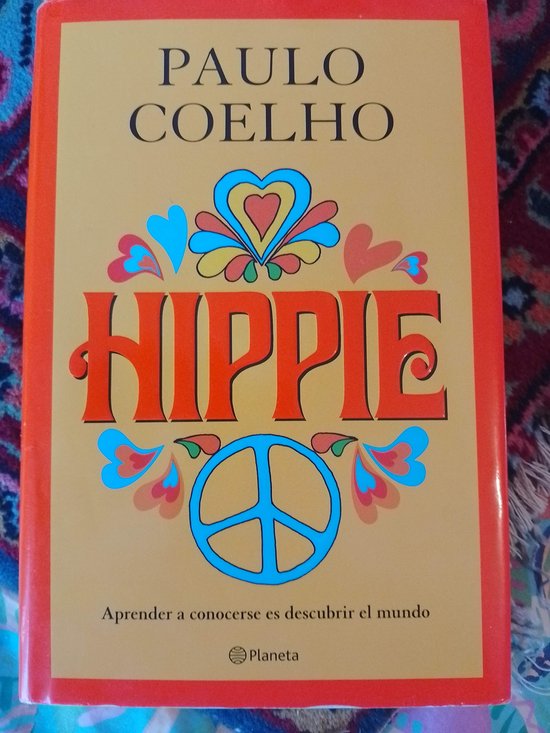 La couverture du livre 'Hippie' de Paulo Coelho est de couleur sable avec des bords orange. Le titre est grand et rouge, avec un symbole de paix et des formes colorées de cœur et de fleur.