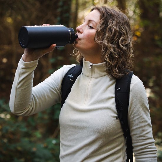 Vrouw met beige NOMAD trui en rugzak, drinkend uit donkerblauwe fles.