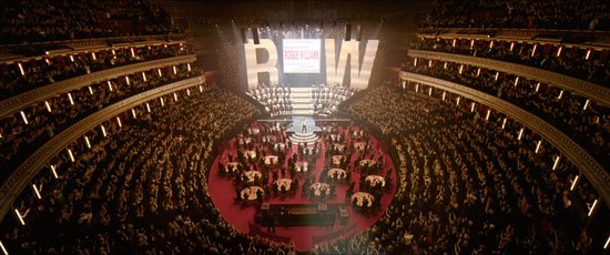 Een artiest staat op een podium voor een groot publiek, met op de achtergrond 'ROBBIE WILLIAMS' verlicht.