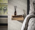 Zwevend nachtkastje van naturel acaciahout met horizontale ribbels en een open lade. Een diffuser en telefoon liggen erop. Naast een bed.