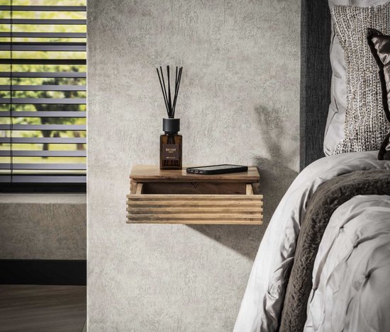 Zwevend nachtkastje van naturel acaciahout met horizontale ribbels en een open lade. Een diffuser en telefoon liggen erop. Naast een bed.