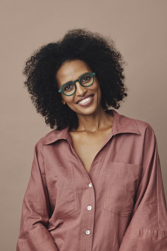 Femme aux cheveux bouclés et chemisier rose, portant des lunettes rondes vertes.