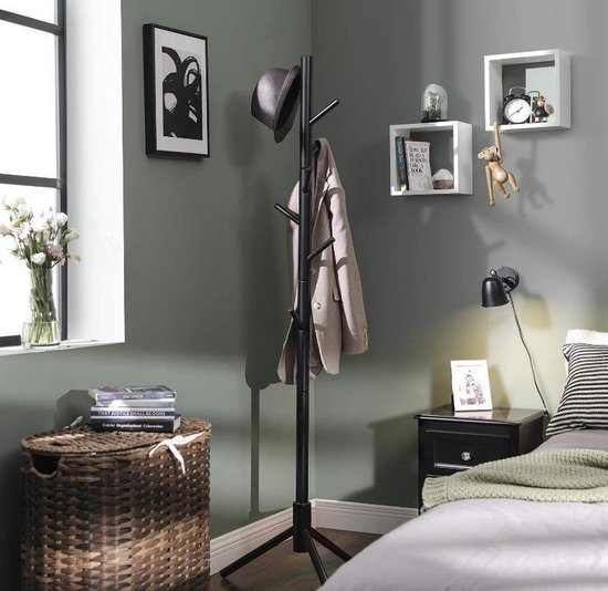 Zwarte staande kapstok met een jas en hoed, in een slaapkamer met een mand, bed en muurdecoratie.