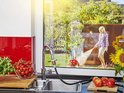 Binnenkraan verbonden met grijze slang; vrouw sproeit planten in tuin.