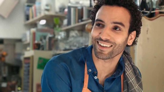 Man met krullend haar in blauw hemd met oranje schort en keukendoek over zijn schouder.
