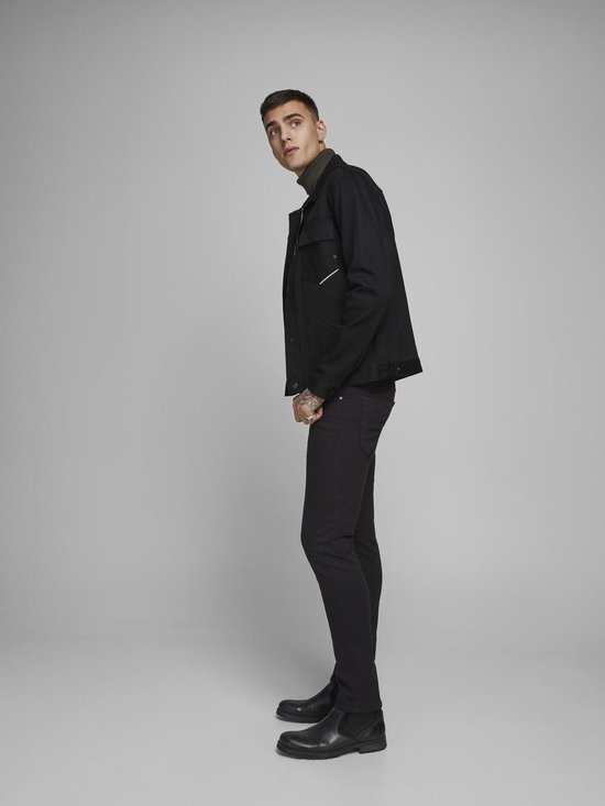 Zij-aanzicht van een model in een zwarte spijkerjack, groene coltrui en zwarte skinny jeans.