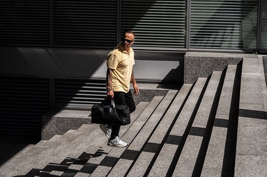Man met geel poloshirt, zwarte jeans en een zwarte sporttas die een trap oploopt.