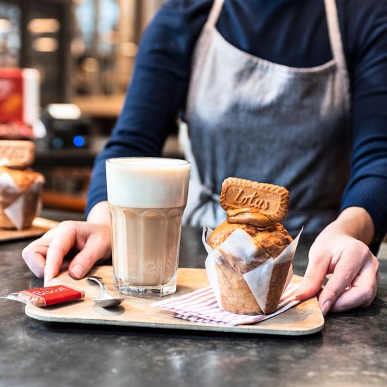 Barista met schort serveert glas met koffie en een muffin met Lotus Biscoff koekje bovenop.