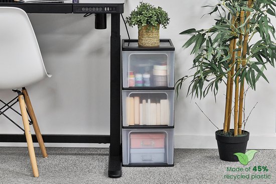 Een zwart ladesysteem met drie transparante lades, naast een bureau met een plant.