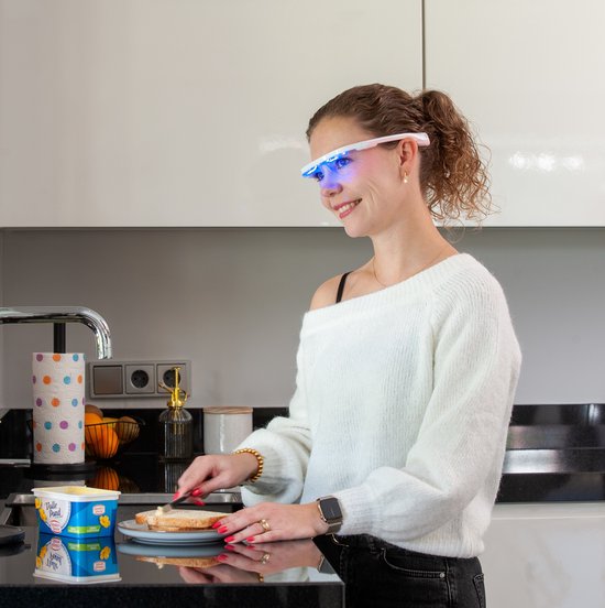 Vrouw met witte AYOlite lichttherapiebril in een keuken, brood besmerend. Blauwe lampjes branden.