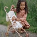 Een kind in roze jurk met golvend haar, zittend bij een roze poppenbuggy met een pop met speen.